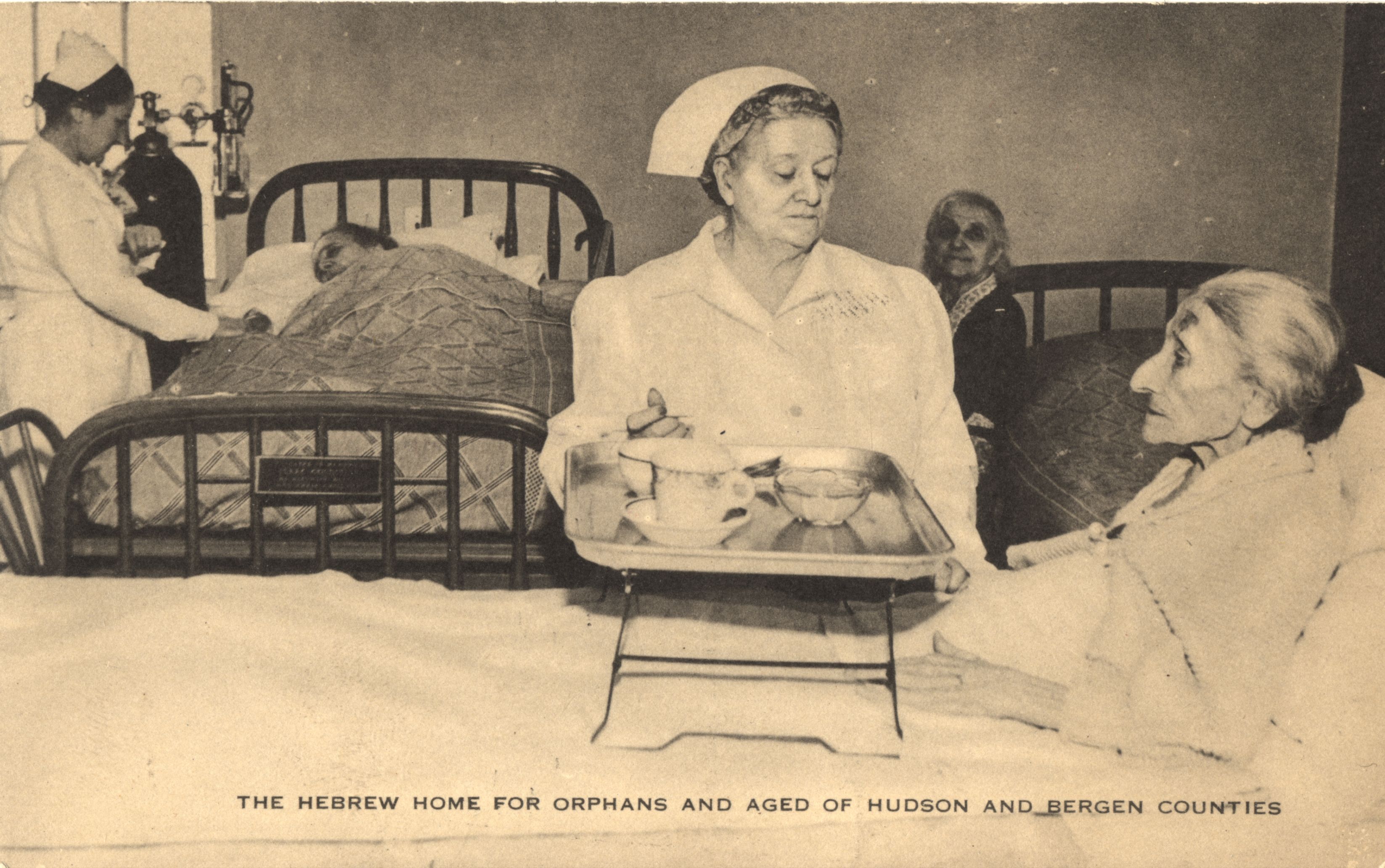 Postcard featuring a black and white photograph of two nurses assisting elderly patients. The nurse in the front is bringing food to an elderly woman.